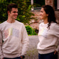 Frau und Mann tragen beide ein cremeweißes Sweatshirt mit AMEN Druck in Regenbogenfarben.